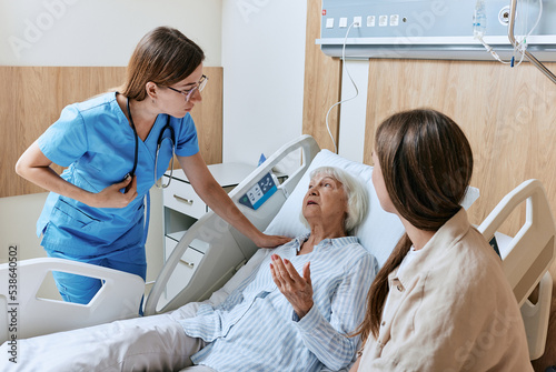 Nurse caring for elderly patient lying in bed of hospital ward, elder care. Daughter supports elderly mother patient during round of ward nurse