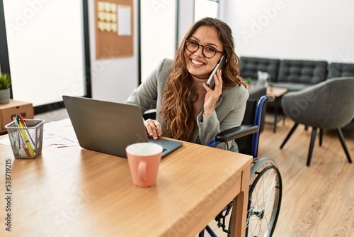 Young beautiful hispanic woman business worker talking on smartphone sitting on wheelchair at office