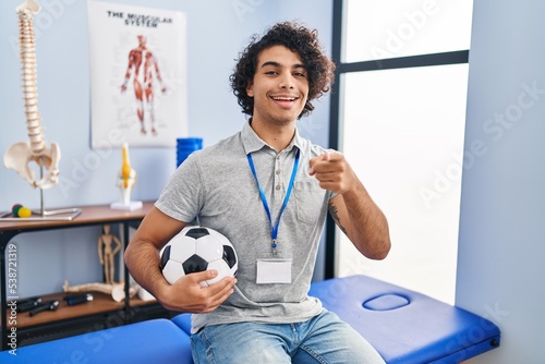 Hispanic man with curly hair working as football physiotherapist pointing to you and the camera with fingers, smiling positive and cheerful