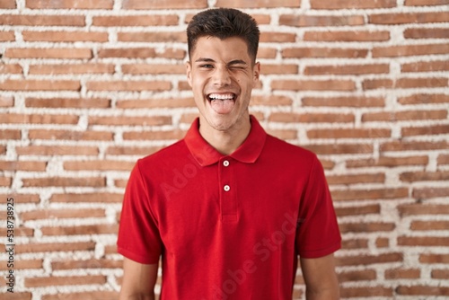 Young hispanic man standing over bricks wall sticking tongue out happy with funny expression. emotion concept.