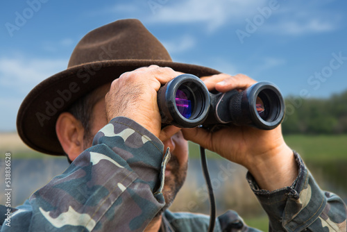 A hunter in a hat with binoculars looks out for prey against the background of the forest.