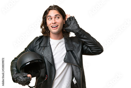 Young handsome man with a motorcycle helmet isolated on green chroma background listening to something by putting hand on the ear