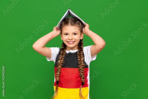 A little girl with the flag of Germany on her T-shirt holds notebooks over her head like a house on a green isolated background.