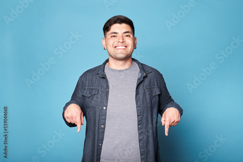 Portrait of a happy latino young man standing, looking at camera, smiling and pointing fingers down on mock up space copy space. Isolated on blue background.