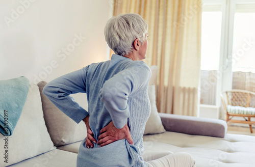 Woman feels back pain, massaging aching muscles. Mature woman feeling morning discomfort in aching back in the living room.