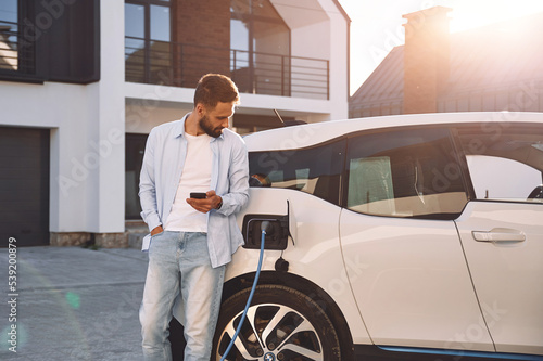 Charging the vehicle near the house. Holding smartphone. Young stylish man is with electric car at daytime