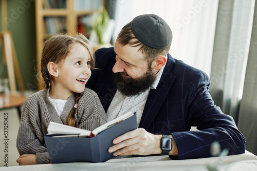 Portrait of smiling jewish father reading book with daughter at home
