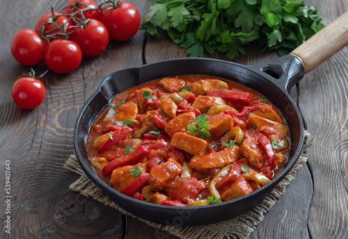 Turkish stew Tavuk sote. Sauteed chicken breast in tomato sauce mixed with onion and colorful bell pepper served in in frying pan. Wooden background, horizontal, selective focus.