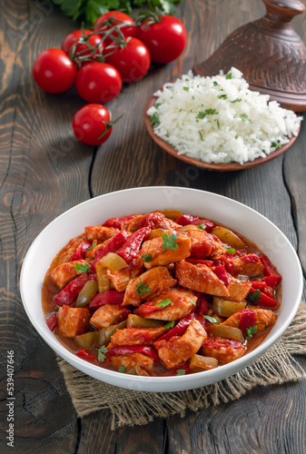 Tomato chicken stew. Braised chicken breast in tomato sauce mixed with onion, red and green bell pepper served with bowl of cooked rice on background. Wooden background, vertical, selective focus.