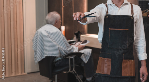 Peluquero irreconocible, mostrando las tijeras, y de fondo cliente senior sentado con la capa, listo para cortar el pelo.