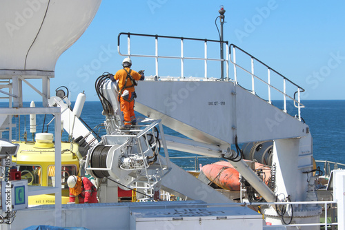 Two seafarers painting ship's crane while out at sea. Both wearing full PPE and fall protection harnesses to prevent fall and injury. Onboard safety culture concept.