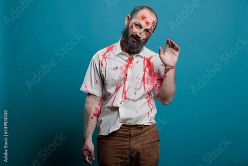 Dangerous zombie waving hello at camera, doing salute gesture over blue background. Creepy apocalyptic evil monster with bloody scars and wounds, brain eating aggressive corpse.