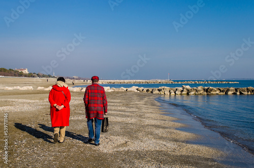 Due persone vestite di rosso camminano lungo la spiaggia del Lido di Venezia in una giornata invernale di sole