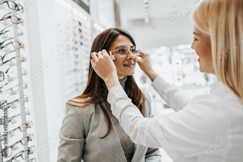 Beautiful and fashionable woman choosing eyeglasses frame in modern optical store. Female seller specialist helps her to make right decision.