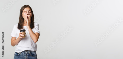 Portrait of tired and sleepy young attractive girl yawning while drinking takeaway coffee, white background