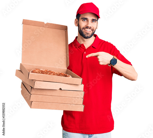 Young handsome man with beard wearing delivery uniform holding boxes with pizza smiling happy pointing with hand and finger