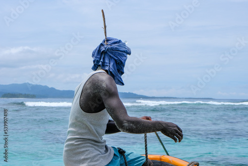 A Melanesian man sitting on the bow of a fishing boat wearing head wrap scarf with a large feather to protect from sun in Bougainville, Papua New Guinea