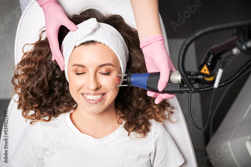 Top view of smiling woman with closed eyes receiving facial skincare treatment in beauty salon. Cosmetologist using erbium ablative laser device while performing resurfacing procedure.