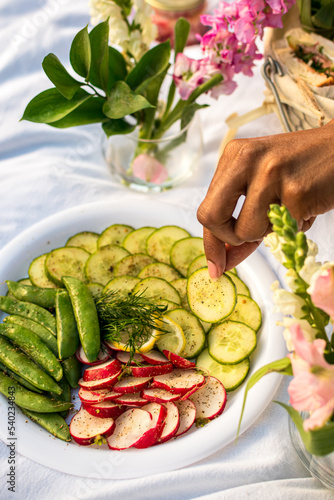 Black woman holding a cucumber slice from crudites platter while enjoying a luxury sunset picnic in the park 