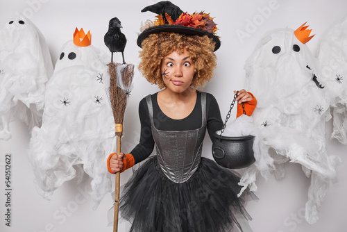 Funny curly haired female wizard with spider on face crosses eyes makes grimace holds broom and cauldron involved in witchcraft wears black hat dress poses around spooky ghosts. Halloween is coming