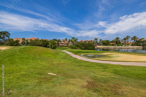 Beautiful view of Aruba's hotel golf course. Green grass and pink buildings behind Green trees on blue sky with white clouds background. 
