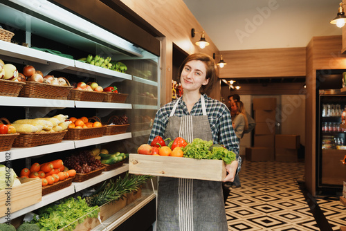 Caucasian young adult saleswoman is carrying a fruit basket and preparing to add stock of fruit to the shelf. Female employee who working in the grocery store looking at the camera with smile face. 