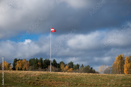 Latvian national flag flutters raised on a tall mast against the background of autumn nature and cloudy sky