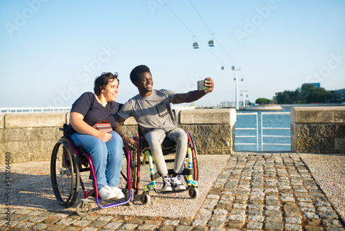 Happy biracial couple taking selfie on embankment. African American man and Caucasian woman in wheelchairs on embankment, holding mobile phone, smiling. Love, relationship, social media concept