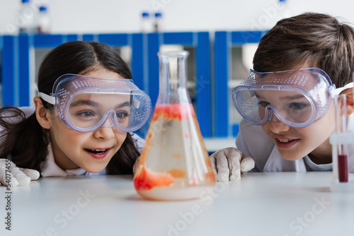 amazed kids in goggles looking at blurred flask with liquid while doing chemical experiment in lab.