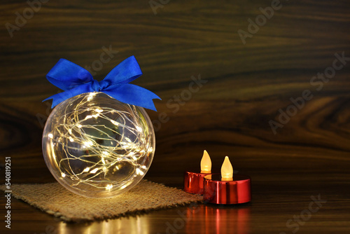 Esfera navideña con luces blancas en el interior sobre un fondo de madera color cafe y velas rojas. Espacio para texto al lado derecho. Concepto Navideño.