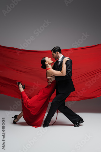 Side view of ballroom dancers performing tango on grey background with red fabric