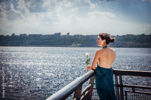 Waiting in the Summer. Young woman with bare back, long blue dress, holding white rose, standing by fence by Hudson River in New York City, looking far away..