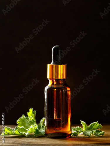 Geranium essential oil in dark glass bottle with fresh scented geranium leaves on old wooden board on black background. Aromatherapy oil, spa massage, mosquito repellent. Copy space, vertical