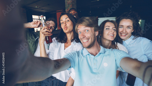 Group of happy colleagues are taking selfie together, young man is holding camera and posing, his coworkers are holding drinks in bottles, laughing and looking at camera.