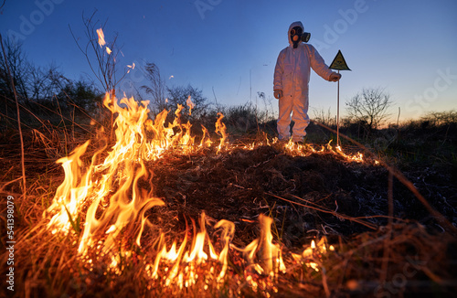 Firefighter ecologist working in field with wildfire at night. Man in suit and gas mask near burning grass with smoke, holding warning sign with skull and crossbones. Natural disaster concept.