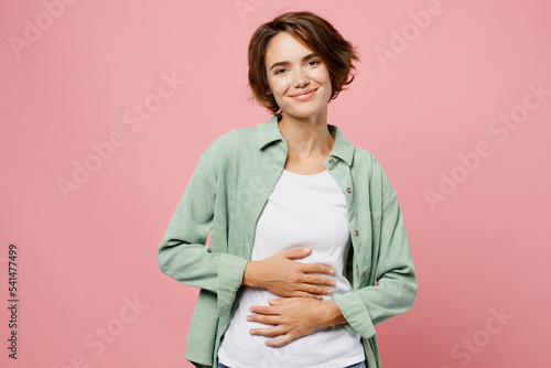 Young smiling cheerful fun happy woman 20s she wear green shirt white t-shirt holds hands on belly look camera isolated on plain pastel light pink background studio portrait. People lifestyle concept.