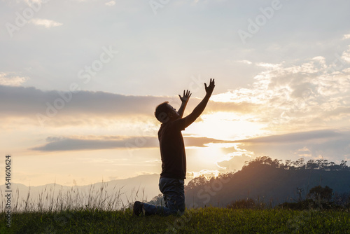 Young male kneeling down with hands open palm up praying to God on the mountain sunset background.