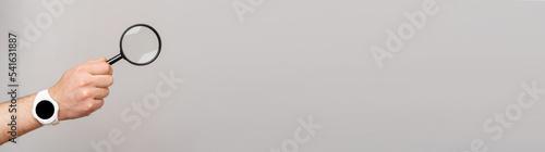 Banner size image of male hand with smartwatch holding magnifying glass over grey background.