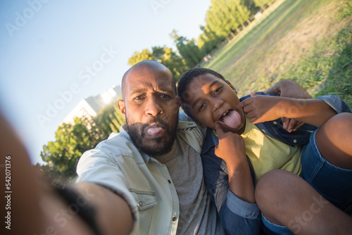 Close-up of man and boy taking funny selfie on field. Bearded dad and his little son sitting on green field showing tongues and both looking at camera. Leisure, childhood and social media concept