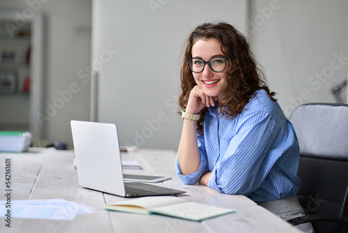 Young happy business woman company employee sitting at desk working on laptop. Smiling female professional entrepreneur worker using computer in corporate modern office looking at camera. Portrait.