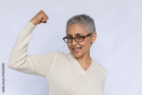 Portrait of happy senior woman showing arm bicep over white background. Confident mature Caucasian woman wearing eyeglasses and white jumper looking at camera and smiling. Pride concept