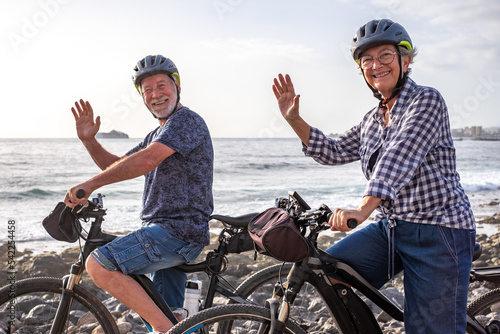 Cheerful senior caucasian couple riding on the pebble beach with electric bicycles waving hands at camera . Authentic elderly retired life concept