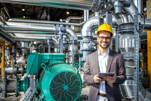 Portrait of power plant engineer standing by gas engines inside energy production factory and checking results on digital tablet.