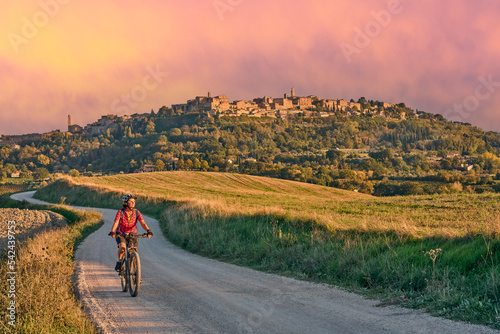 nice senior woman riding her electric mountain bike between olive trees in the Ghianti area with medieval city of Montepulciano in background, Tuscany , Italy