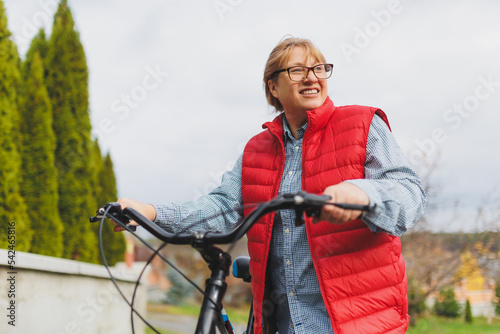 Middle aged smiling mature woman holdingt a bike with her hands on the grass on a green field. Summer or Autumn Country Vacation and Adventure Concept