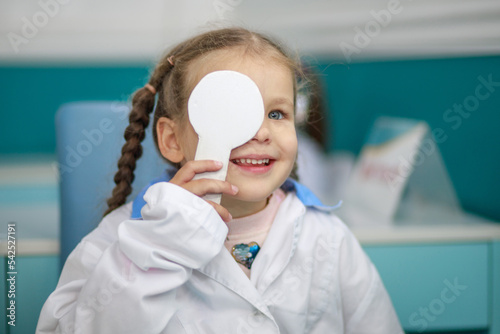 Happy girl plays ophthalmologist doctor, child closes one eye with a white flap to check vision, children's role-playing games. 