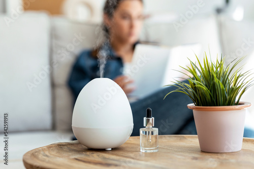 Shot of essential oil aroma diffuser humidifier diffusing water articles in the air while woman reading a book sitting on coach.