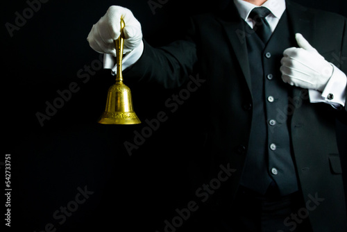 Portrait of Butler or Waiter in Dark Suit and White Gloves Holding Gold Bell on Black Background. Service Industry and Professional Hospitality.