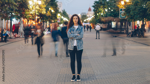 Portrait of stylish young lady tired of usual haste standing in the street among whizzing people and looking at camera. Time, youth and modern society concept.