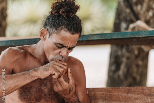 Imagen horizontal de un hombre adulto de cabello rizado sin camisa en la playa encendiendo su pipa para fumar hierba. 